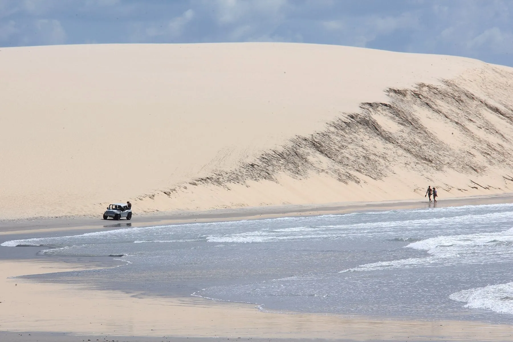Veículo 4x4 atravessando a areia ao pé de uma grande duna à beira-mar